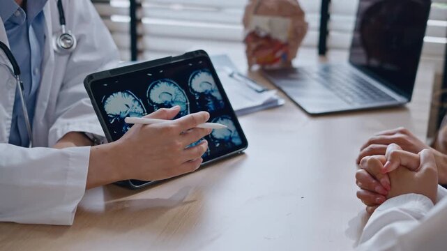 Professional Asian male neurologist doctor explaining brain MRI scan results on digital tablet to a Couple - relationship in modern clinic. Concept of healthcare, diagnosis, and medical technology.