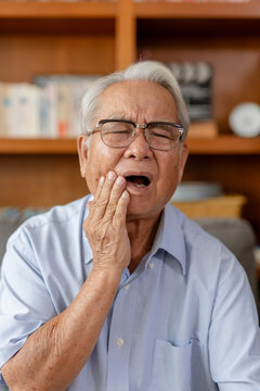 Senior man feeling severe toothache pain holding hand to jaw with distressed expression sitting indoors experiencing dental problem