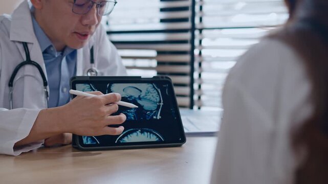 Professional Asian male neurologist doctor explaining brain MRI scan results on digital tablet to a Couple - relationship in modern clinic. Concept of healthcare, diagnosis, and medical technology.