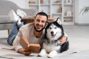 Handsome young man lying on floor and reading book with cute husky dog at home © Pixel-Shot