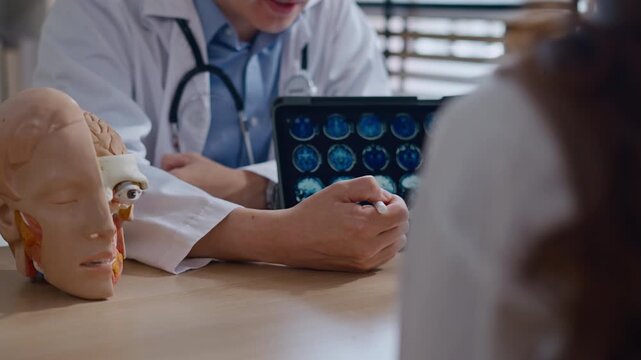 Professional Asian male neurologist doctor explaining brain MRI scan results on digital tablet to a Couple - relationship in modern clinic. Concept of healthcare, diagnosis, and medical technology.