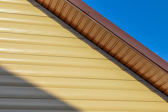 Beige vinyl siding covers the exterior facade of a house underneath a brown metal roof against a clear blue sky. Quality exterior cladding protects the building walls from harsh weather conditions whi