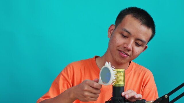 Asian guy tapping and scraping plastic hairbrush on golden microphone capsule against turquoise backdrop