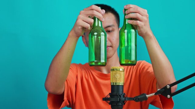 Asian guy tapping green beer bottles in front of  golden microphone on cyan background 