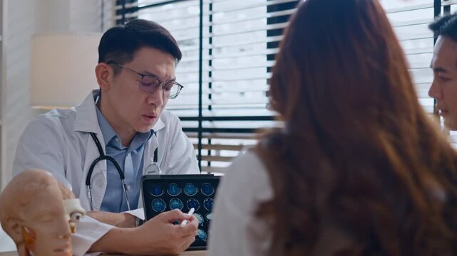 Professional Asian male neurologist doctor explaining brain MRI scan results on digital tablet to a Couple - relationship in modern clinic. Concept of healthcare, diagnosis, and medical technology.