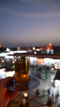 Marrakech, morocco. August 9, 2026. A hand holding a traditional glass of mint tea above the illuminated jemaa el fnaa square in marrakech, morocco, during a warm evening