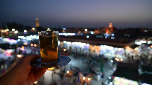 Marrakech, morocco. August 9, 2026. Hand holding a traditional moroccan mint tea in a glass against the beautifully blurred background of the jemaa el fnaa square in marrakech