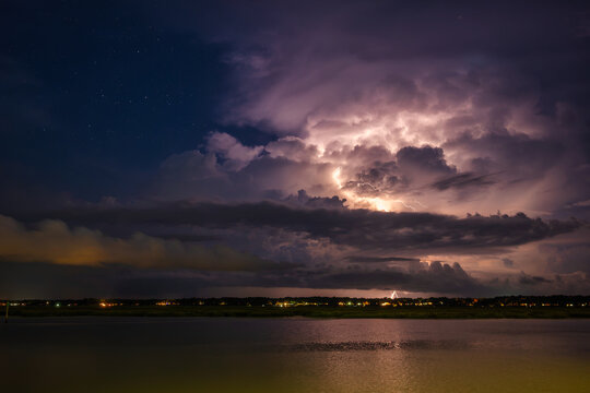 Lightning illuminates clouds over a river at night, showing a stormy sky and distant city lights