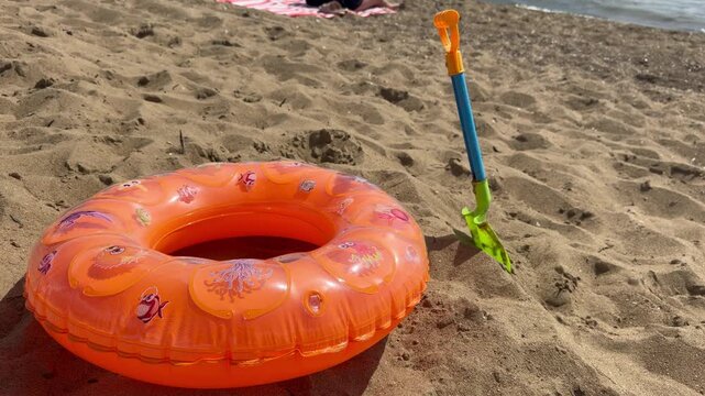 Inflatable orange swimming ring and colorful beach toys resting on warm sand by the sea, sunny summer vacation scene, concept of tourism, family leisure, beach recreation