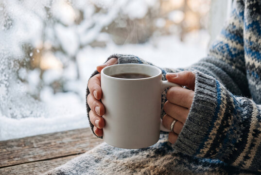 Woman's hands in a patterned knit sweater hold a white mug. A frosty, snow-covered window with trees is seen in the background. Cozy winter scene.