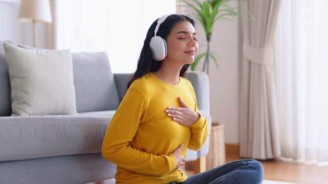 Asian woman meditates and practices Zen meditation at home, focusing on breathing exercises and seeking spiritual peace in her living room. She sits on the floor, listening podcast to headphones