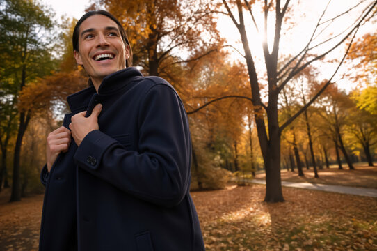 Smiling young man wearing a navy coat in an autumn park. Vertical portrait of a happy male enjoying a sunny fall day outdoors. Seasonal fashion and lifestyle concept