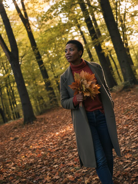 Black woman holding a bouquet of autumn leaves in a forest. Vertical portrait of a female walking in the woods during fall. Seasonal outdoor lifestyle and nature concept