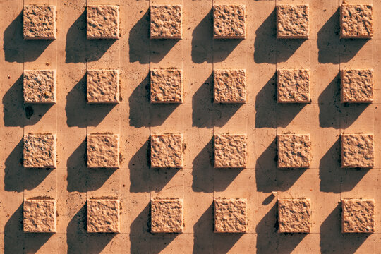 Top-down texture of tactile paving tiles with raised square studs casting strong diagonal shadows in warm sunlight.