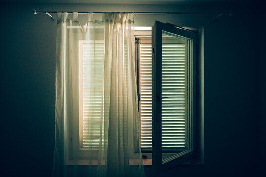 Interior shot from a dim room looking toward an open window with white curtains, where bright exterior daylight filters in through horizontal shutter louvers.