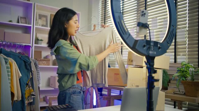 Young Asian woman fashion entrepreneur live streaming selling clothes online with smartphone ring light and laptop in home studio, representing small business owner and professional women.