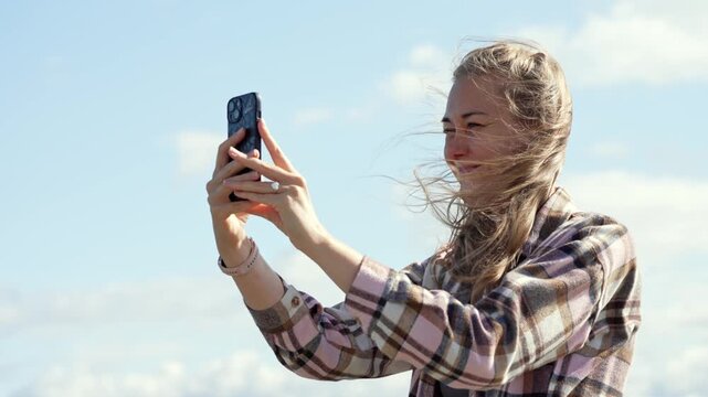 Radiant woman with windblown hair stands outdoors during a bright midday to take photos with her mobile device while enjoying the warmth of the sun and clear weather in a peaceful natural environment.