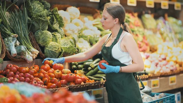 Shop seller put goods on display case. Girl creates stock of tomato in display case, forms showcase. Vegetable shop near house. Store with long rows of display cases, customers waiting 