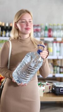 Girl buyers examine goods, selects large capacity bottle of water in store. Visitor make choice of assortment, choose and buy water in plastic bottle. Customer read label, examine customer information