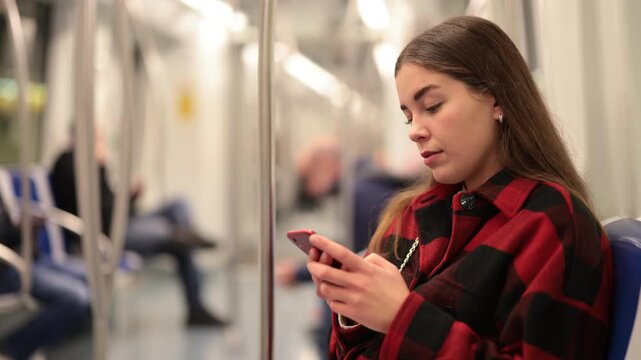 Young European woman is sitting in a subway car and typing a message on her mobile phone. Girl using online navigator to use public transport 