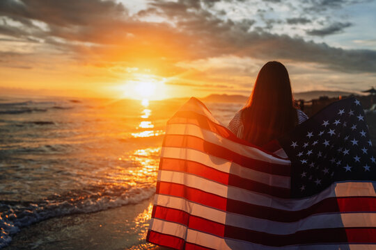 Beautiful patriotic young woman with the American flag held in her standing in the summer sunset