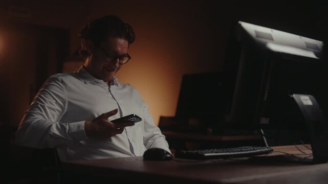 Cheerful young entrepreneur in dark office taking vulgar crude photo on mobile phone. Happy smiling businessman texting on smartphone and laughing procrastinating during late night work, slow motion.