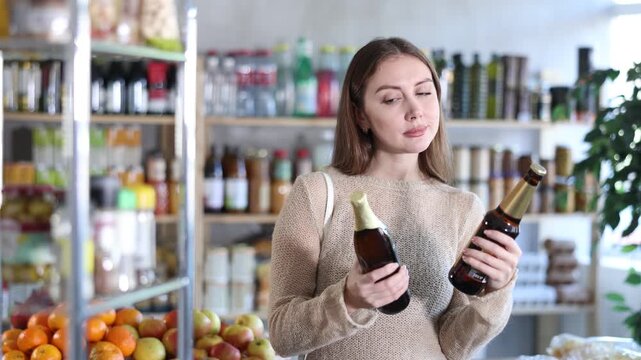 Girl hold bottles, choose and buy light alcohol drink in store. Market visitor consider and make choice, buy beer. Client hesitates before buying, choose beer, reads labels on bottles 