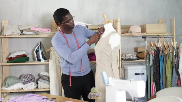 African American man tailor connects parts of dress on mannequin. Female craftsman attaches front part of dress to back with pins. 