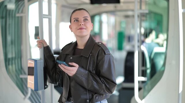 Young woman stands inside moving tram, holding smartphone while commuting through city 
