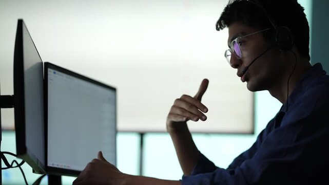 A young engineer standing with a tablet raises a hand as he presents to the room, three lit screens standing behind his shoulder at an AI company in Bangkok, Thailand.
