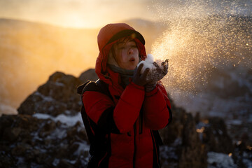 Happy woman in a warm down jacket blowing snow from her palms among snowy mountain rocks at golden hour, enjoying a playful winter adventure. © Sonya Monshtein