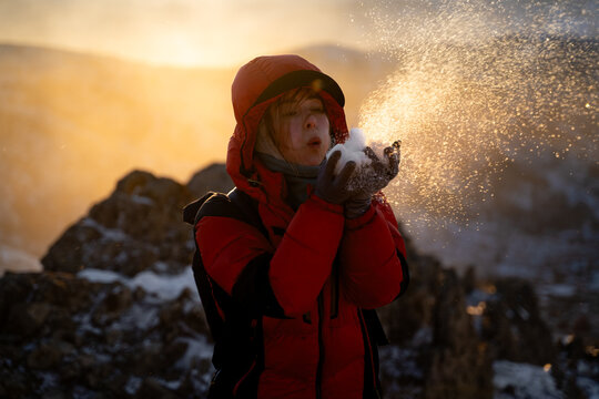 Happy woman in a warm down jacket blowing snow from her palms among snowy mountain rocks at golden hour, enjoying a playful winter adventure.
