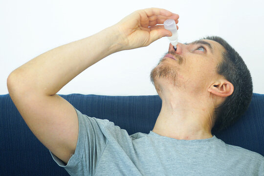 Man reclining on sofa applying nasal drops from small bottle. Conveys cold treatment routine, respiratory health recovery, at-home symptom relief, and self-care during seasonal respiratory infection.