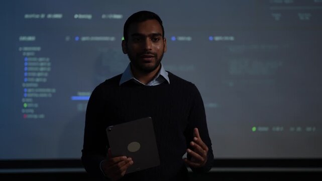 A machine learning engineer in a dark knit sweater talks straight to camera with both hands on a tablet while a live agent execution log scrolls on the wall screen in a dark tech office.