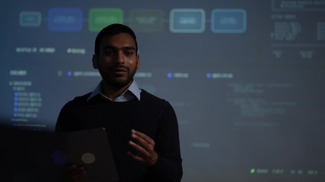 A young software engineer holding a tablet walks through a node graph of AI agent pipeline stages glowing on the projection screen behind his shoulder in a dark tech office.