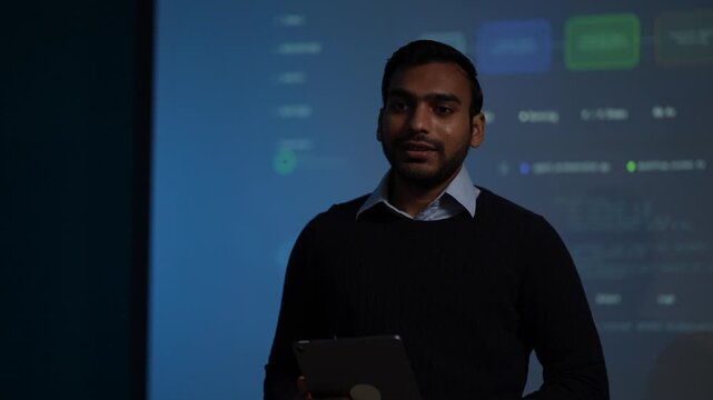 A confident AI developer in a navy sweater gestures out to the side and then smiles as a blue agent monitoring interface fills the screen behind him in a dark tech office.