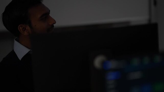 A bearded developer seen in tight profile glances sideways at his monitors in a nearly dark engineering room lit only by the screens at an AI company in Bangkok, Thailand.