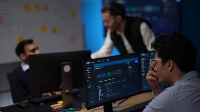 Three developers spread across a row of workstations review dashboards together while a colleague in a black vest leans in over the desk to join the discussion in a dark tech office.