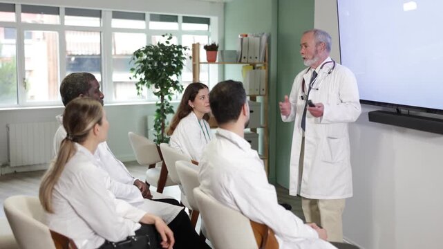 During lesson, mature male doctor with remote controller tell material, speaks to group of students. Lecturer leads class in audience, explains material to audience, provide practical cases 