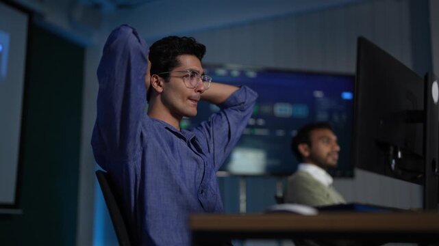 A tired developer at a late night desk stretches both arms behind his head, then drops back to the keyboard to keep working on the model inside a dark AI startup office.
