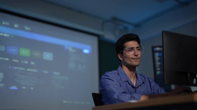 A software engineer lit by a projected dashboard breaks into a small satisfied smile before turning back to the code running on his own display in a dark tech office.