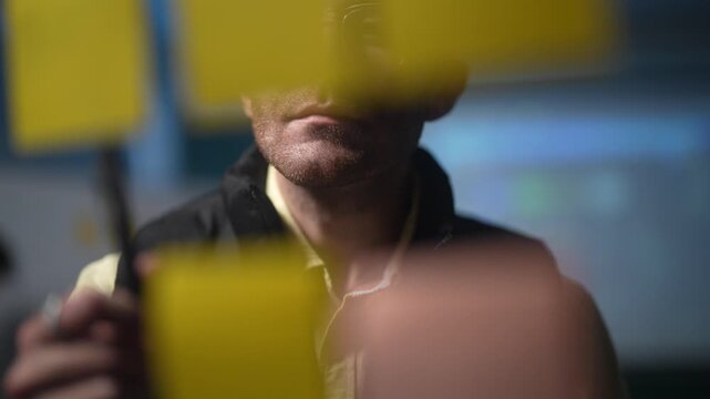 A bespectacled team member seen through a note covered pane adds another sticky to the board and studies where the new idea belongs in the plan in a dark tech office.