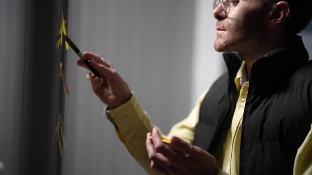 A designer with a marker in his hand gestures at the sprint board and then reaches up to hang one more note on the glass wall inside a dimly lit machine learning startup office.