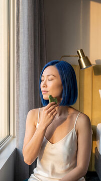 Serene woman using jade gua sha tool by sunlit window at home