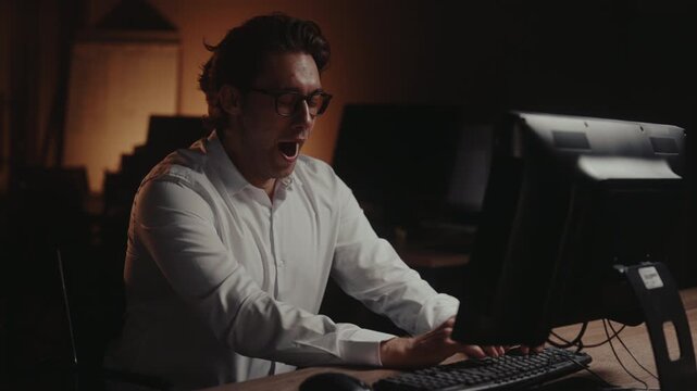 Man working late in dark office feels tired and overwhelmed, yawning, struggles to stay awake, finally falls asleep at computer keyboard. Concept of burnout and deadline pressure, slow motion.