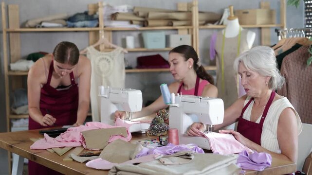 Mature seamstress and a young assistant are sewing clothes next to a fashion designer who is drawing sketches and patterns on a tablet. Dressmaking team is working . High quality 4k footage