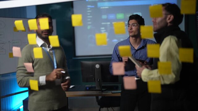 Three teammates on both sides of a glass note wall argue a workflow through, one reaching up to reposition a note while the others watch it land in a dark tech office.