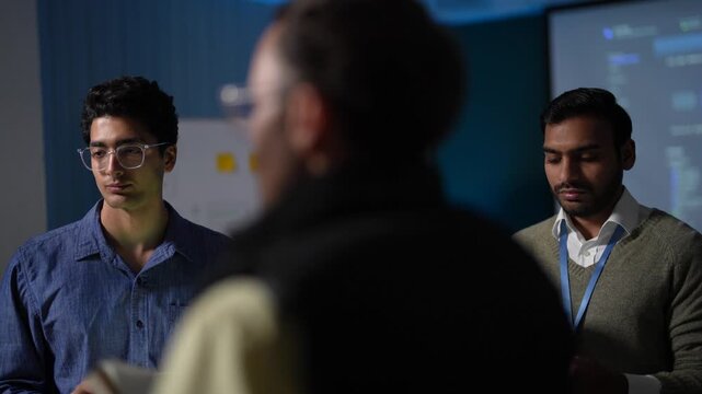 Two engineers facing a colleague with his back to camera listen and answer during a standing review, one of them holding a tablet at his chest in a dark tech office.