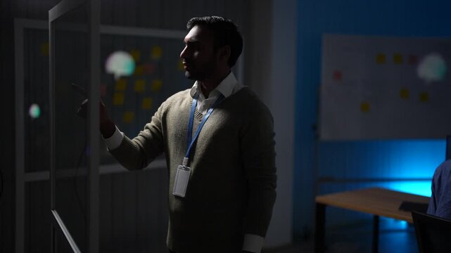 A team member in an olive sweater steps up to the glass wall and points out the note that breaks the rest of the sequence inside a dimly lit machine learning startup office.