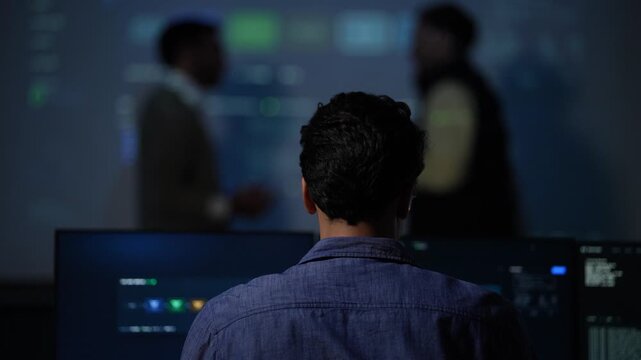 The back of a seated developer's head fills the frame while two blurred colleagues discuss the pipeline on the screen ahead of him inside a dark AI startup office.